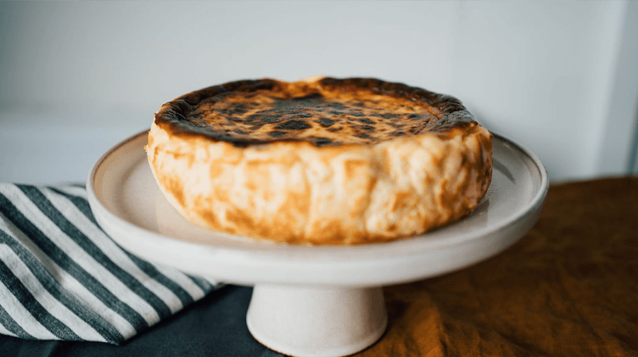 La Maritxxu Basque cheesecake on a cake stand with a stripy towel and white wall as background