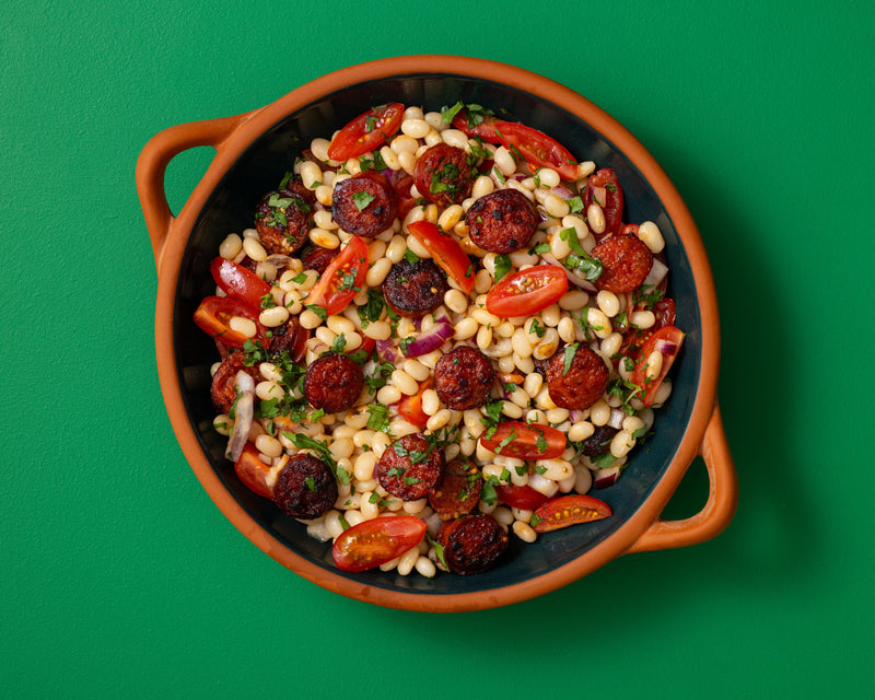 A top-down view of a terracotta bowl filled with a colourful salad containing white beans, red tomatoes, chorizo, and herbs, set against a solid green background.