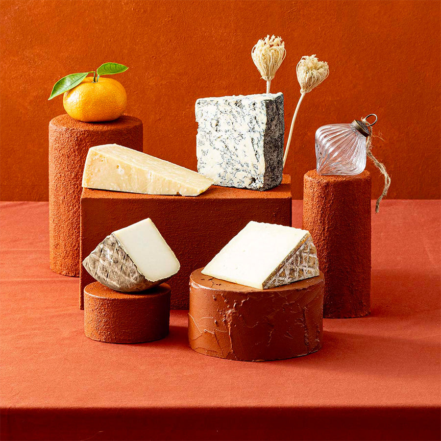 An assortment of Spanish cheeses displayed on terracotta-coloured pedestals against a warm terracotta background. The cheeses include various textures and styles.