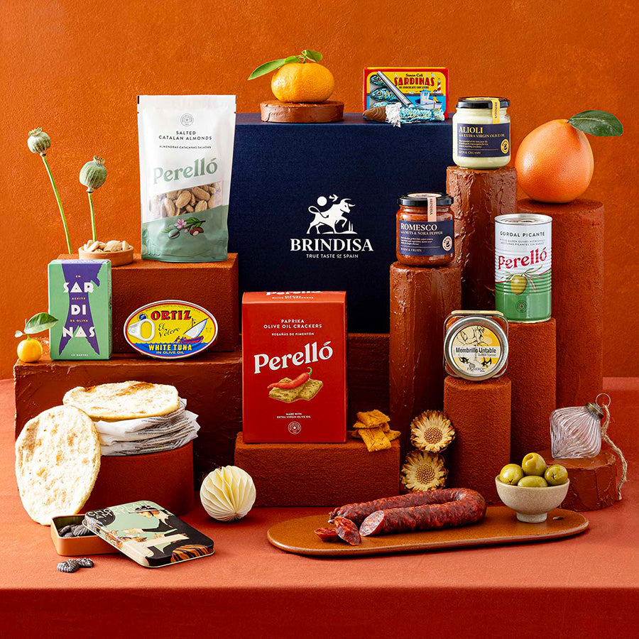 A styled gift assortment of Spanish food products, arranged on terracotta-coloured pedestals, set against a warm terracotta background.