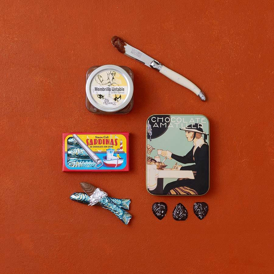 A styled flat-lay of Spanish food products: a jar of membrillo, a tin of chocolate leaves and a box of chocolate sardines.