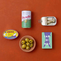 A styled flat-lay of food items on a terracotta background: a can of Perello gordal picante, a tin of sardines, an Ortiz white tuna tin and a bowl of green olives.