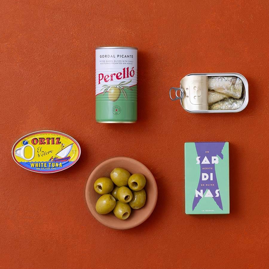 A styled flat-lay of food items on a terracotta background: a can of Perello gordal picante, a tin of sardines, an Ortiz white tuna tin and a bowl of green olives.