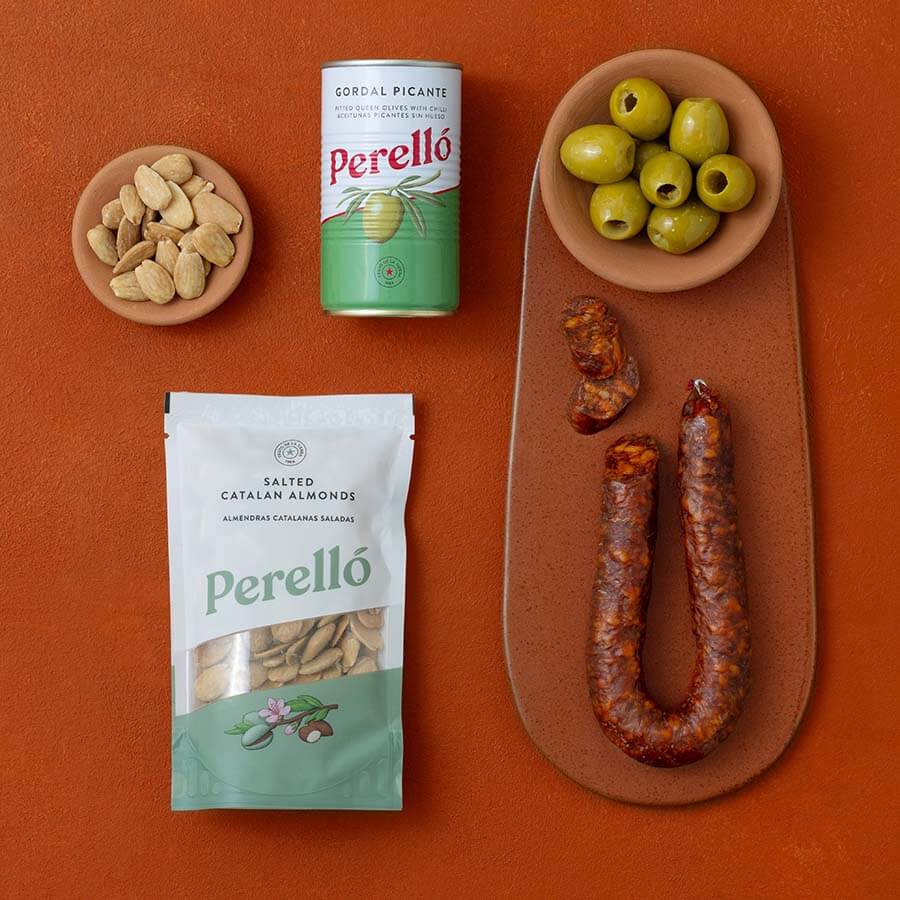 A styled flat-lay of Spanish food products on a terracotta background: Perello olives, Perello Catalan almonds and chorizo.