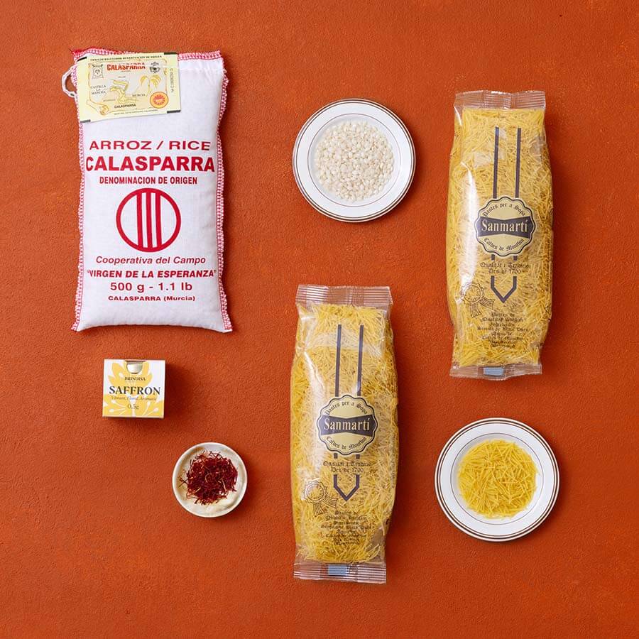 A styled flat-lay of food products on a terracotta background, featuring a white bag of Calasparrara rice, two packages of Sanmarti fideus, a small box of Saffron, and two small bowls containing saffron and fideus.