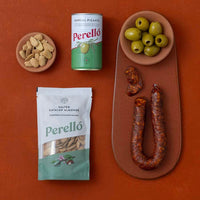 A styled flat-lay of Spanish food products on a terracotta background: Perello olives, Perello Catalan almonds and chorizo.
