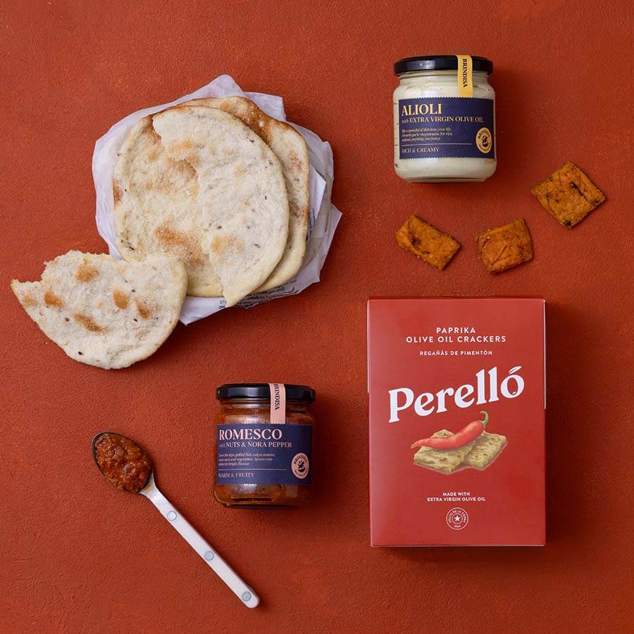 A styled flat-lay of Spanish food products on a terracotta background: a basket of flatbread, a jar of Alioli, a jar of Romesco, a red box of Perelló olive oil crackers, and Tortas de Gavino.