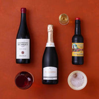 Three bottles of wine and a bottle of sparkling wine arranged on a terracotta background, with two glasses and an open bottle cap nearby.
