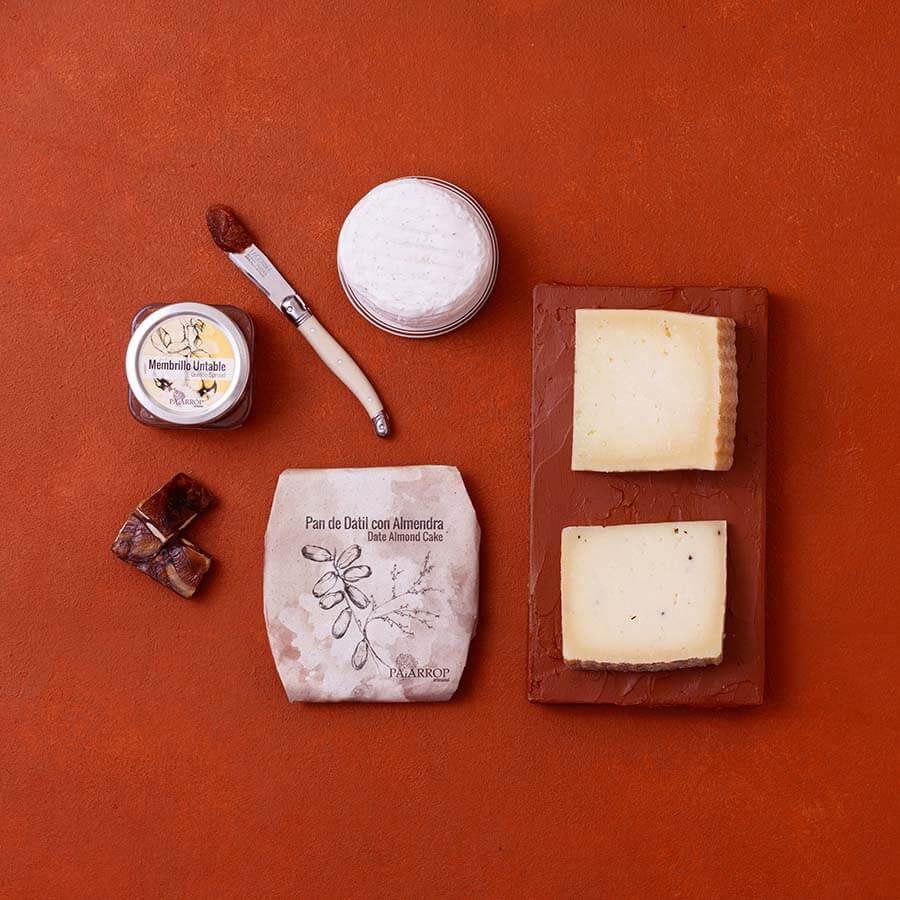 An overhead flat-lay of artisanal Spanish cheeses and membrillo on a terracotta background.