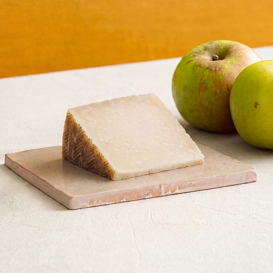 A wedge of Manchego cheese on a rectangular stone serving board, with two green apples behind it on a light surface against a warm yellow-orange background.