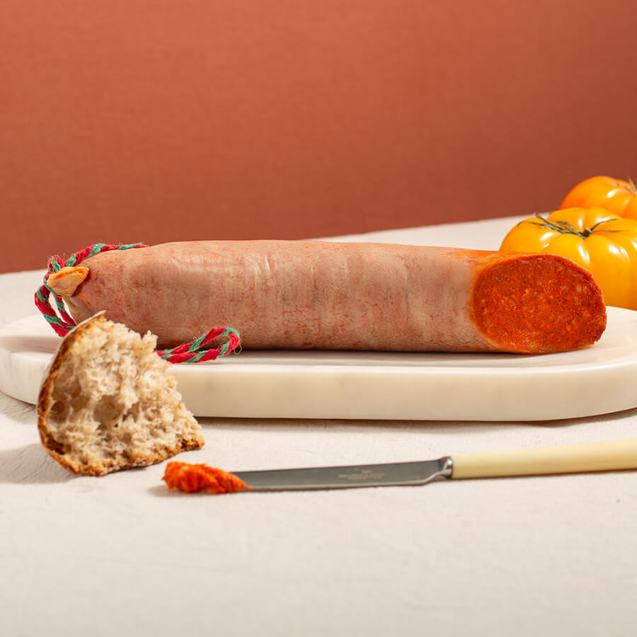 sobrasada from mallorca, spreadable Spanish sausage on a wood board. There's yellow tomatoes at back and, a bit of bread and one knife at the front with a bit of the sausage paste