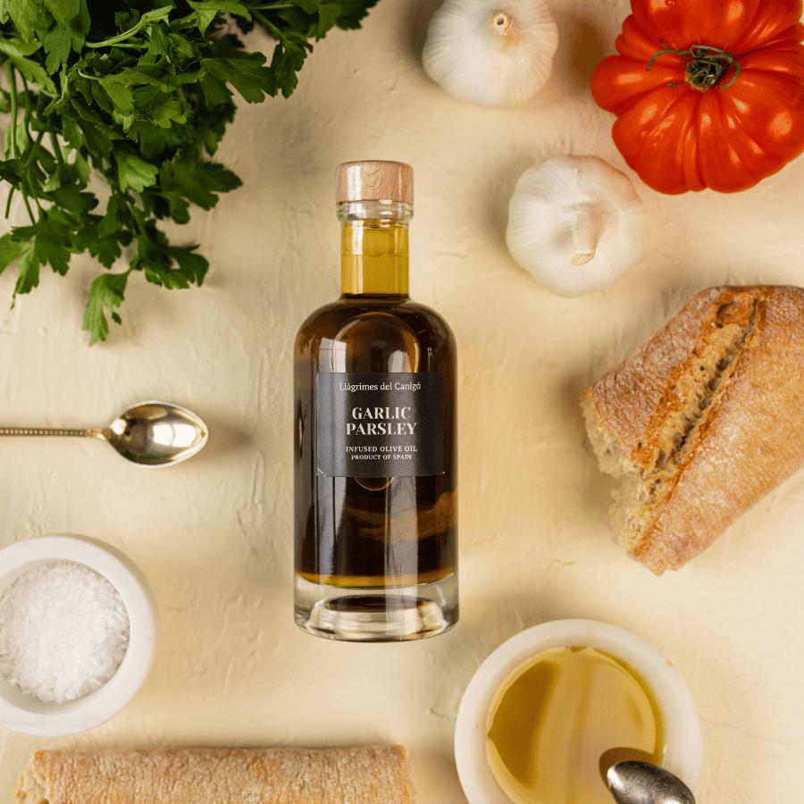 Bottle of garlic and parsley infused olive oil laid flat on a table and surrounded by bread, salt, parsley, tomato and garlic.