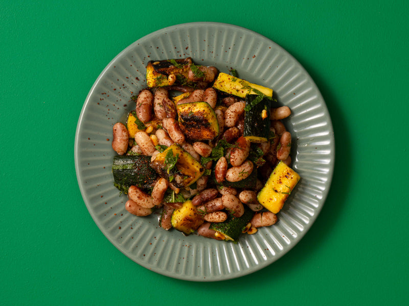 A top-down view of a light gray ceramic plate filled with beans and courgettes, set against a solid green background.