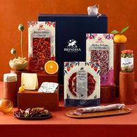 A Spanish gourmet food gift set arranged on a terracotta surface against a matching terracotta background. The set includes a large navy Brindisa box, packaged cured meats, cheeses and accompaniments.