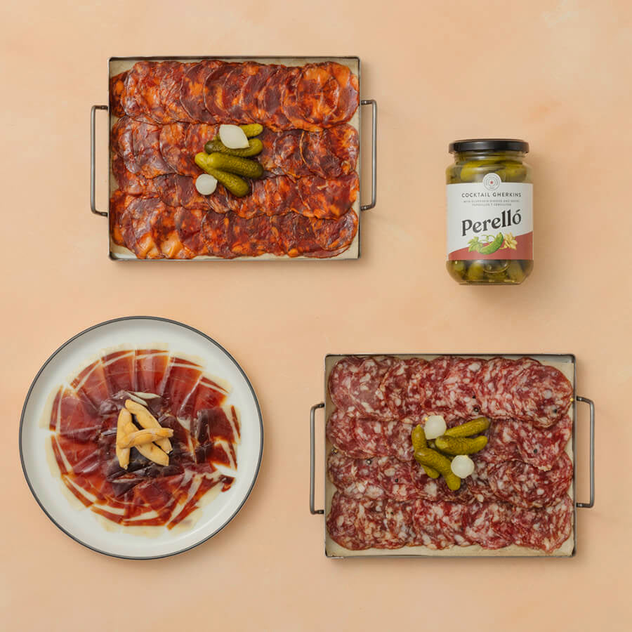 Assorted cured meats with pickles and a jar of Perello pickles on a beige background