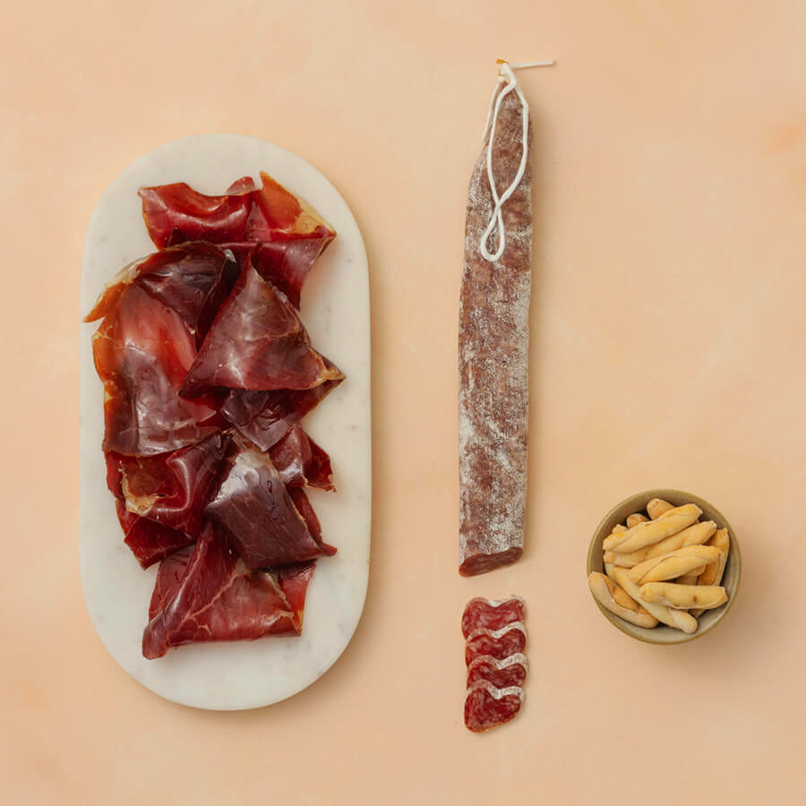 Platter of sliced cured meat with a whole cured meat product and a bowl of bread sticks on a beige background