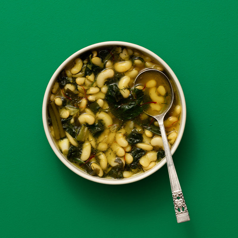 A top-down view of a white bowl filled with a soup containing white beans, dark green leafy vegetables, and oil, with a silver spoon resting inside, set against a solid green background.