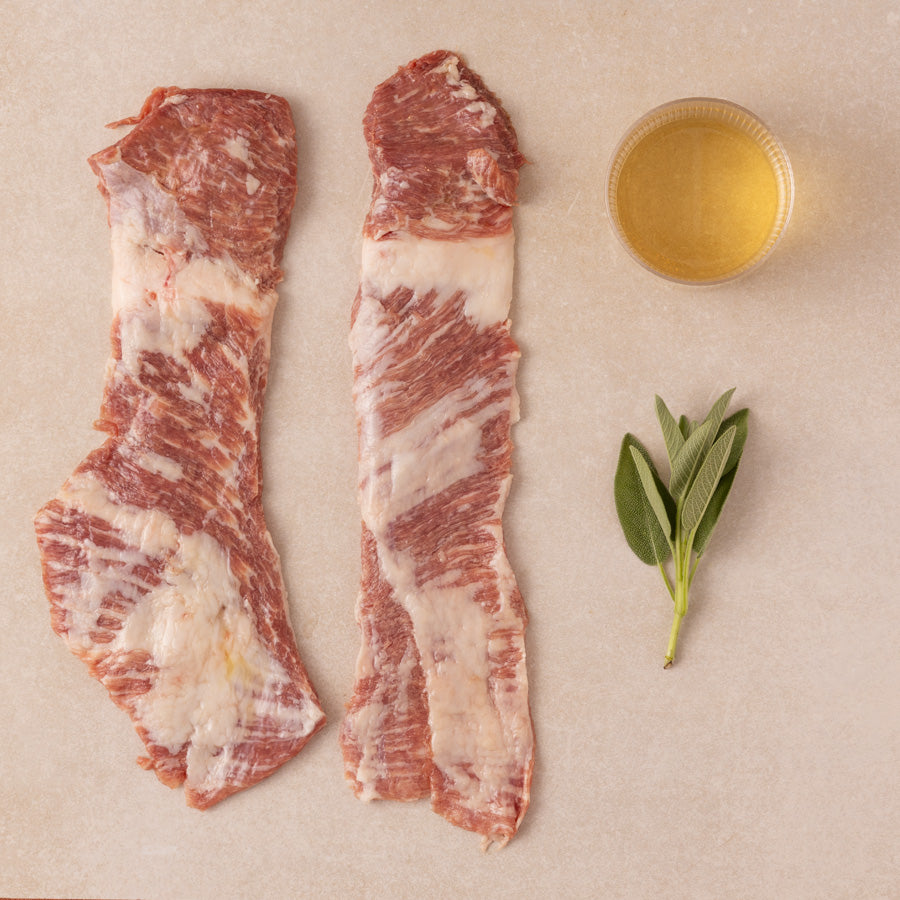 Two raw meat slices (Iberico Secreto) with a small bowl of white wine and sage leaves on a beige surface