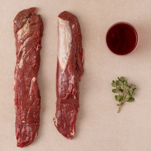 Two pieces of raw meat (iberico solomillo) with a small bowl of red wine and a sprig of thyme on a beige surface.