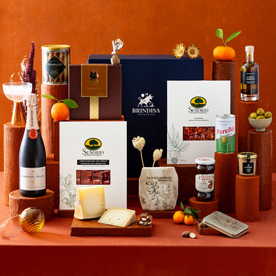 A styled gourmet gift hamper featuring an assortment of Spanish products including cheese, charcuterie, sparkling wine and snacks, arranged on terracotta pedestals against a matching terracotta background.