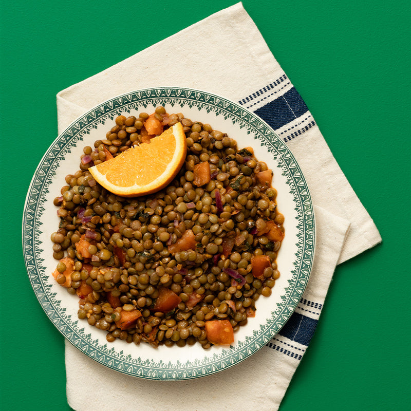A top-down view of a plate of cooked lentils with a lemon wedge, served on a patterned green and white ceramic plate over a cream napkin with a blue stripe, set against a solid green background.