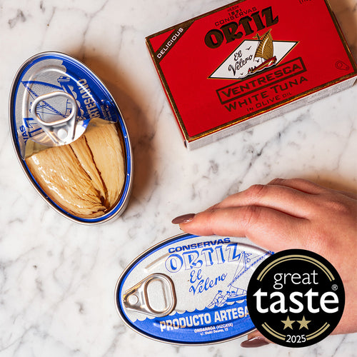 Cans of Ortiz Ventresca white tuna on a marble surface with a hand holding one can, and a Great Taste award badge.