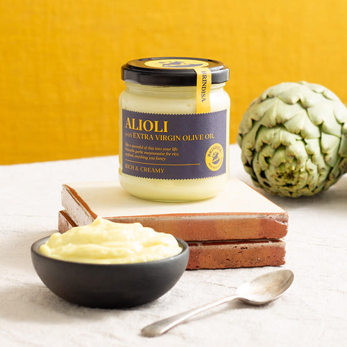 Jar of Brindisa Alioli with extra virgin olive oil beside a fresh artichoke, ceramic tiles, and a bowl of creamy alioli with a spoon