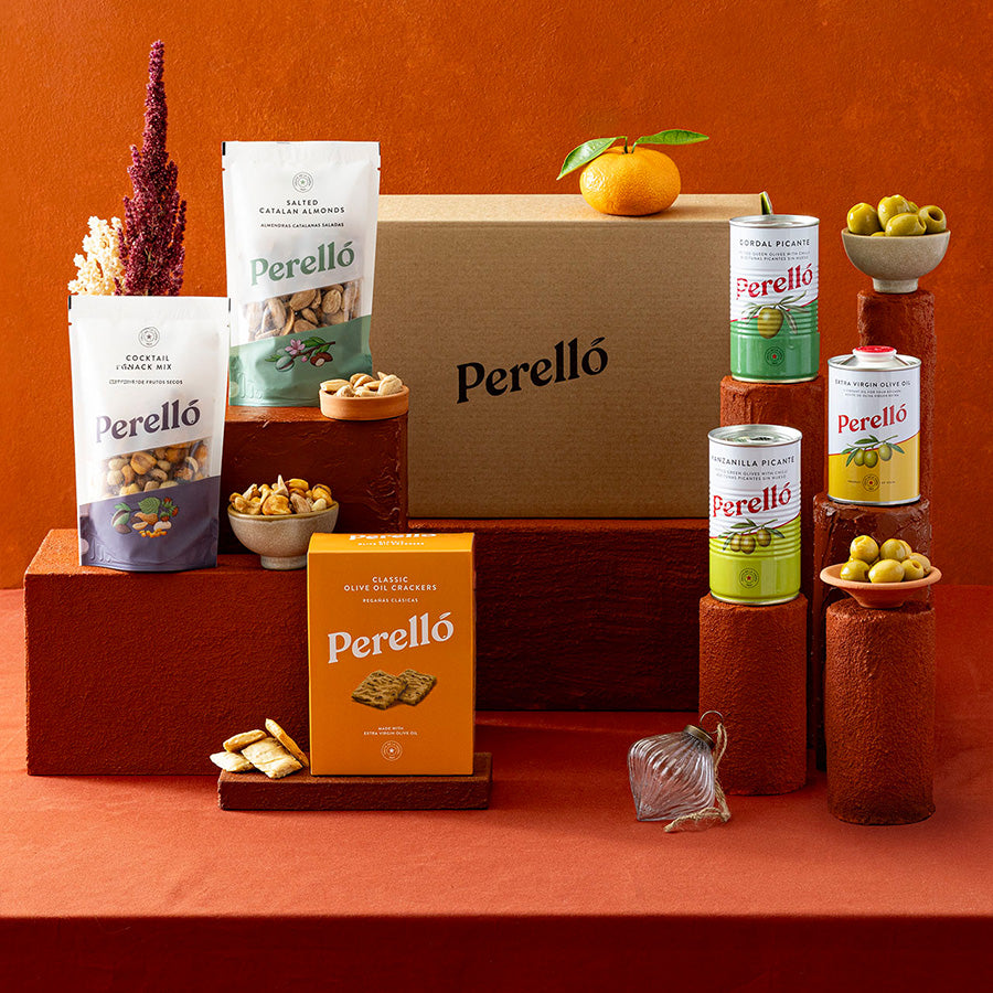 A Perelló snack assortment display on a terracotta background, featuring a large brown cardboard box, two bags of mixed nuts, a box of crackers, tins of olives and arranged on wooden pedestals with small bowls of olives and dried flowers.