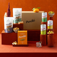 A Perelló snack assortment display on a terracotta background, featuring a large brown cardboard box, two bags of mixed nuts, a box of crackers, tins of olives and arranged on wooden pedestals with small bowls of olives and dried flowers.