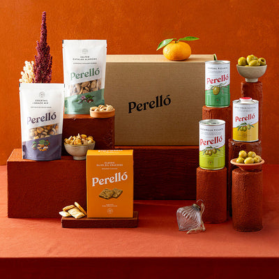 A Perelló snack assortment display on a terracotta background, featuring a large brown cardboard box, two bags of mixed nuts, a box of crackers, tins of olives and arranged on wooden pedestals with small bowls of olives and dried flowers.