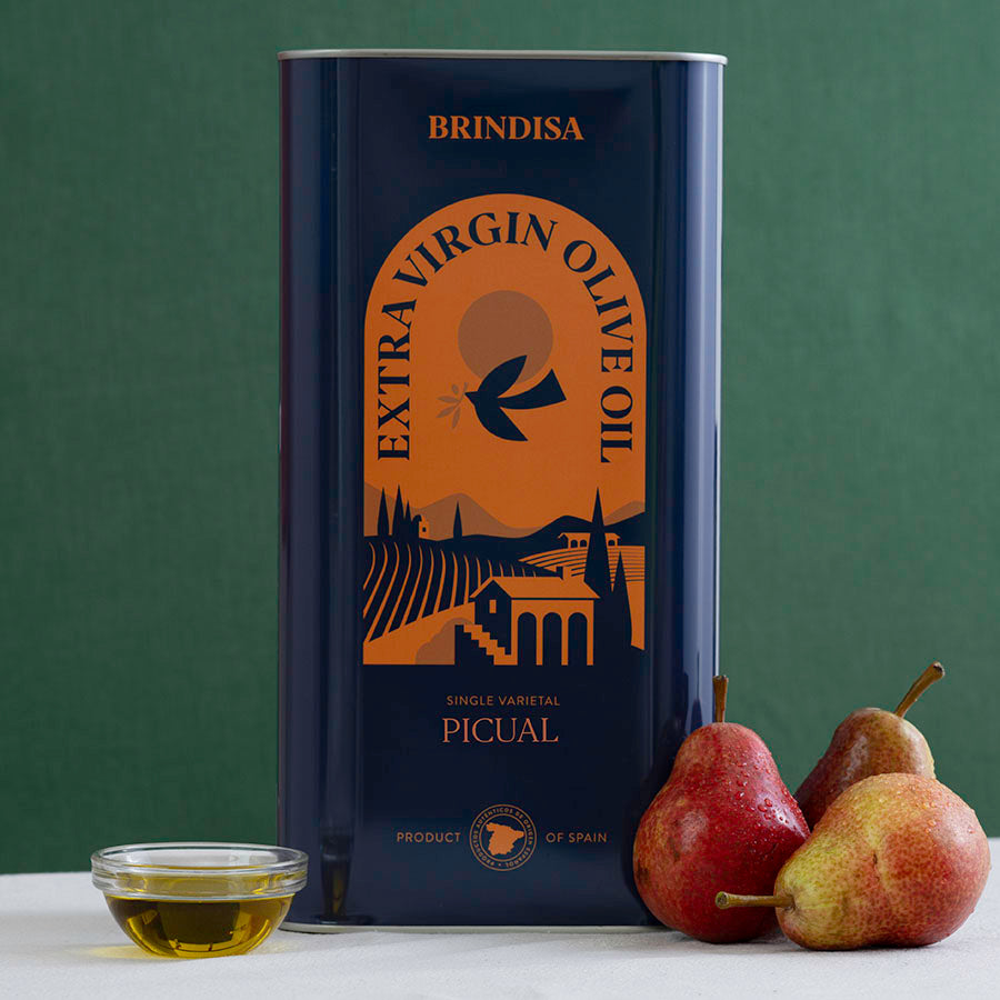 A tall tin of Brindisa Extra Virgin Olive Oil Picual stands on a white surface against a dark green background, with a small glass bowl of olive oil and three pears placed in front of the tin.