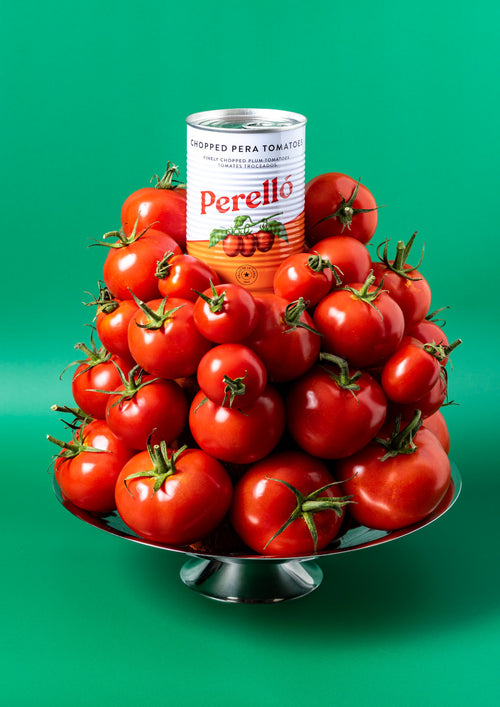 A tower of fresh tomatoes with a tin of Perello chopped pera tomatoes on top, placed on a metal plate against a green-blue background.