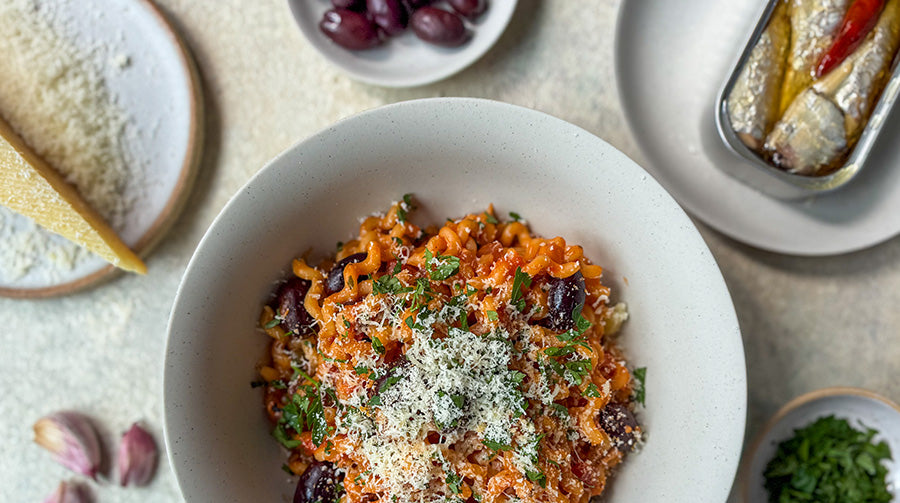 Sardine puttanesca in a pasta bowl with lots of parmesan cheese on top and the ingredients displayed around 