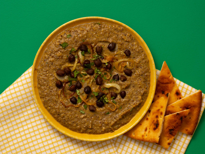 A yellow bowl filled with a thick brown dip, topped with chickpeas and caramelised onions, served on a yellow-and-white napkin with triangular pieces of bread on a green background.