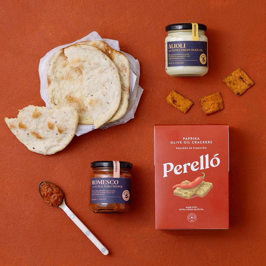 A styled flat-lay of Spanish food products on a terracotta background: a basket of flatbread, a jar of Alioli, a jar of Romesco, a red box of Perelló olive oil crackers, and Tortas de Gavino. 