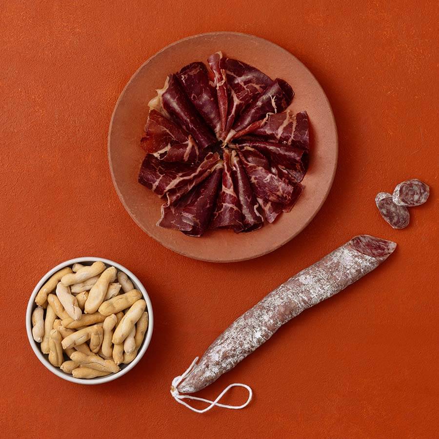 A styled flat-lay of cured meats and picos camperos on a terracotta background.