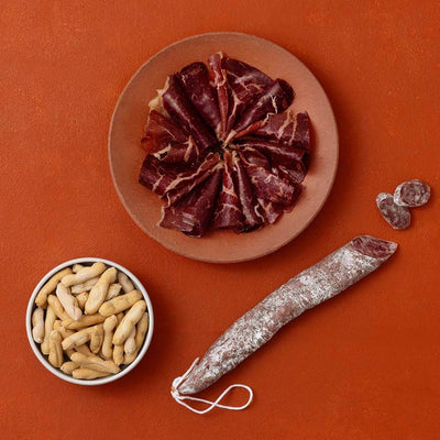 A styled flat-lay of cured meats and picos camperos on a terracotta background.