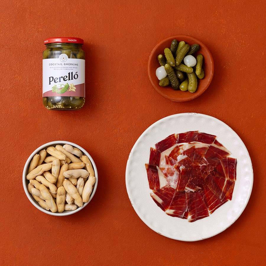 A styled flat-lay of Perello cocktail gherkins on a textured terracotta background, accompanied by a bowl of sliced cured ham, a bowl of pickled onions, and a bowl of picos camperos.