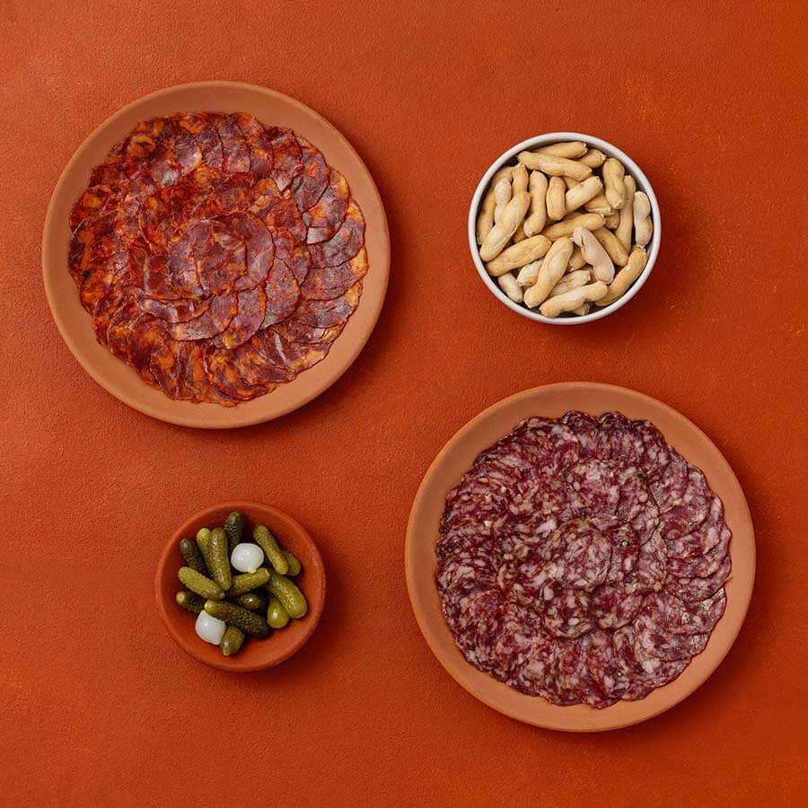 Three terracotta plates and a small white bowl arranged on a warm orange-brown background, containing sliced cured meats, pickles and picos camperos.