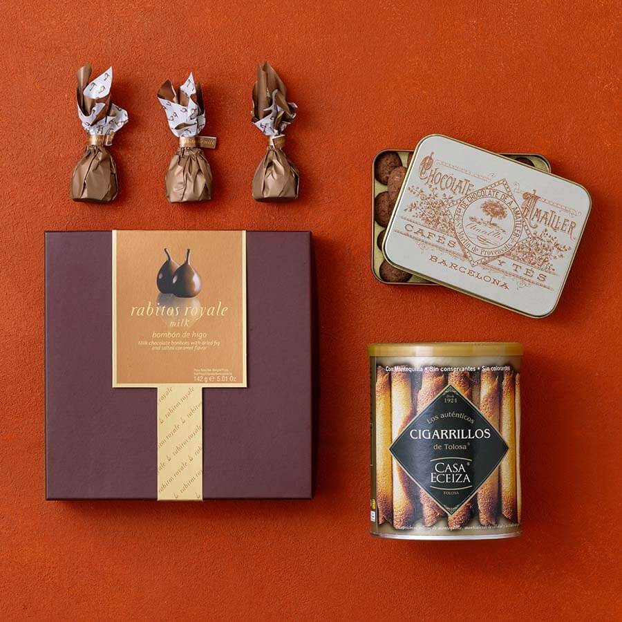 A curated flat-lay of gourmet Spanish snacks and chocolates on a terracotta background.