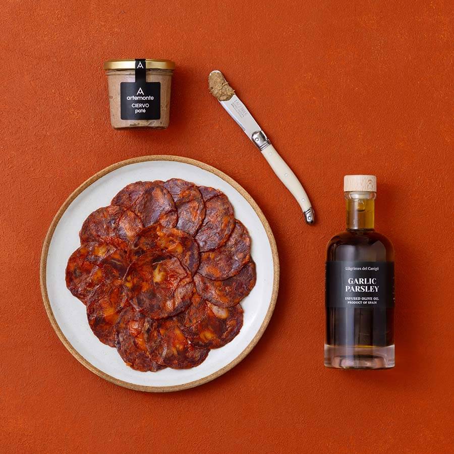 An overhead flat-lay of Spanish foods, including sliced chorizo on a plate, a jar of pate, and a bottle of flavoured olive oil.