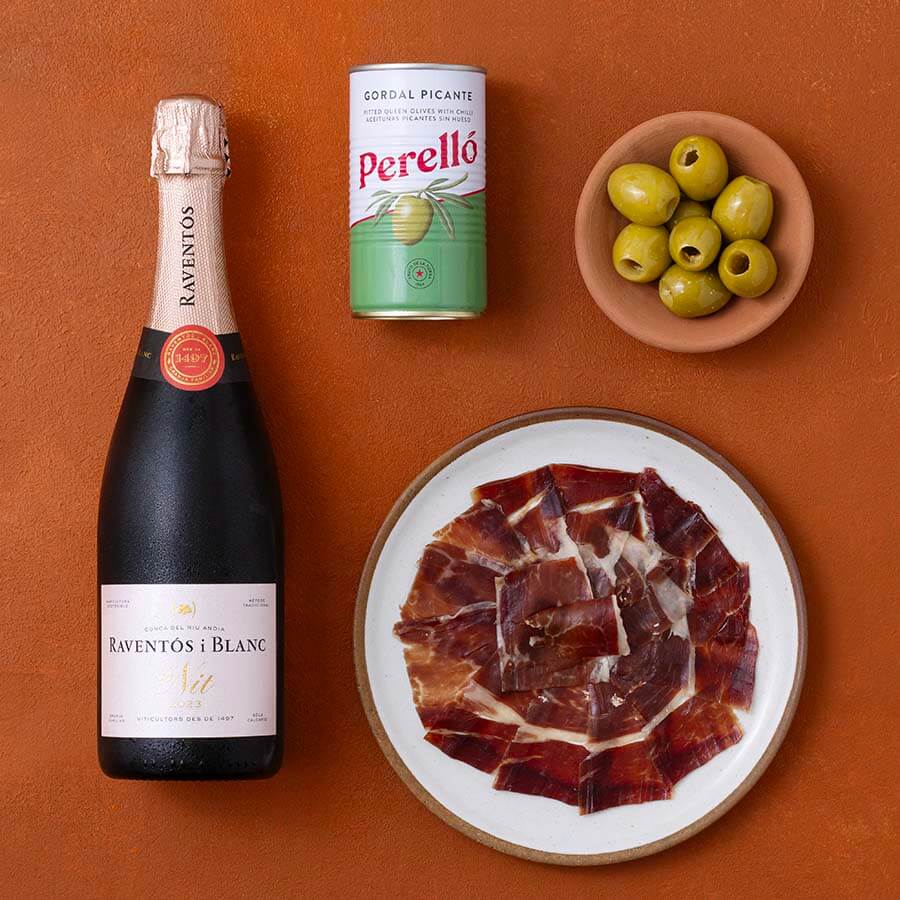 A styled flat-lay of a bottle of Raventos Blanc sparkling wine, a can of Perello Gordal Picante, a small bowl of green olives, and a plate of sliced cured ham on a terracotta background.