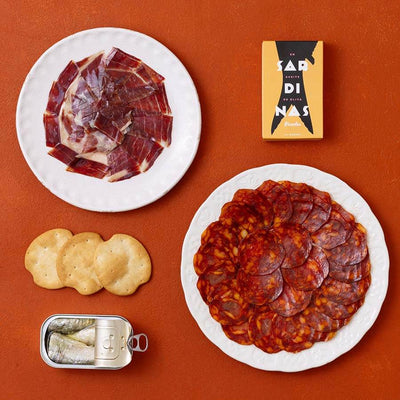 A styled flat-lay of Spanish charcuterie items on a terracotta background: a white plate with sliced cured ham, a white plate with sliced chorizo, a small open tin of sardines and three round crackers.