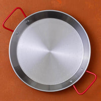 A round stainless steel paella pan with a perforated center and bright red side handles, shown from a top-down angle on a textured terracotta background.