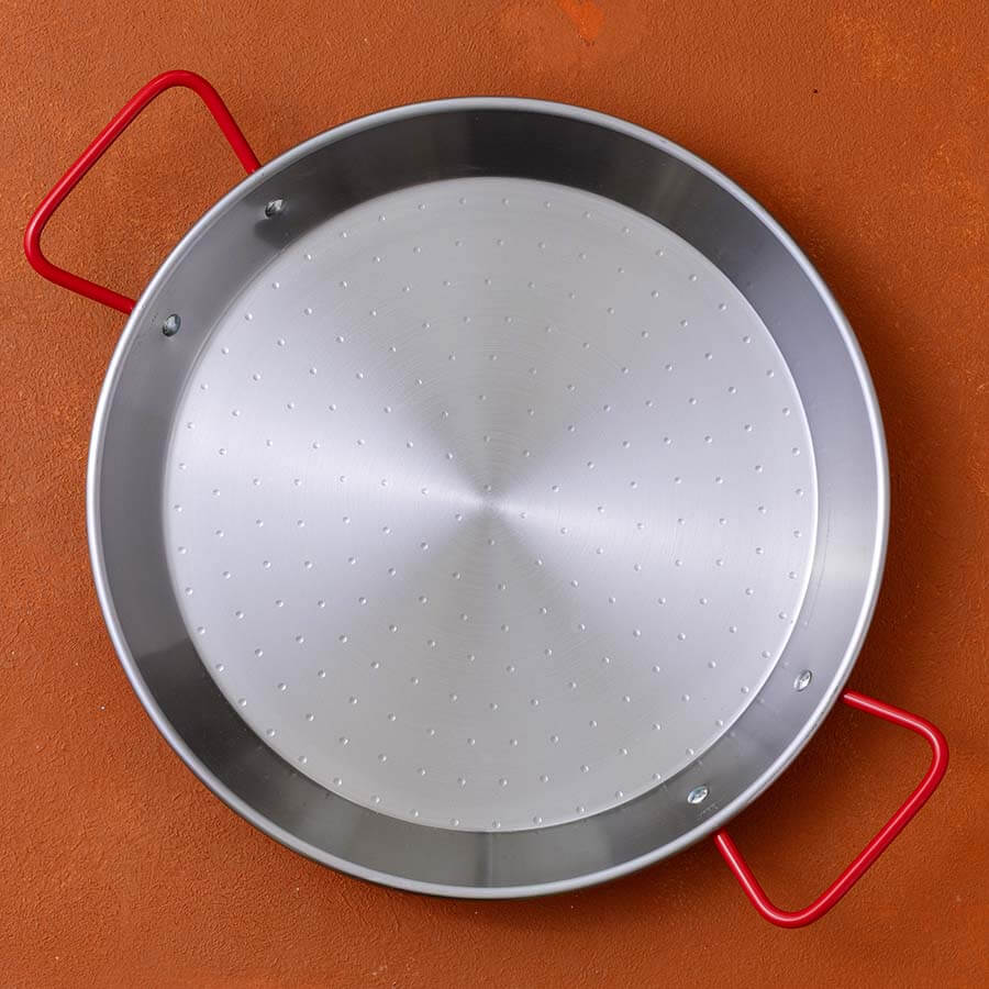 A round stainless steel paella pan with a perforated center and bright red side handles, shown from a top-down angle on a textured terracotta background.