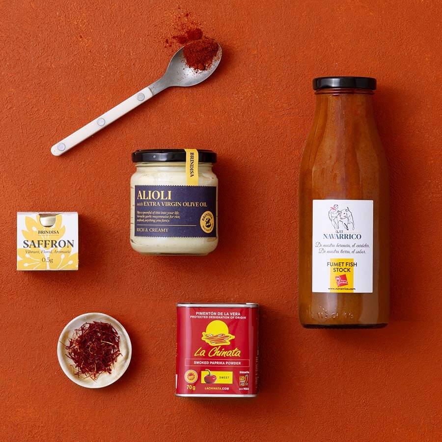 A styled flat-lay of multiple food products on a terracotta background: a large bottle of fumet, a jar of alioli, a box of saffron, a can of La Chinata paprika, a small dish with saffron, and a spoon with paprika. 