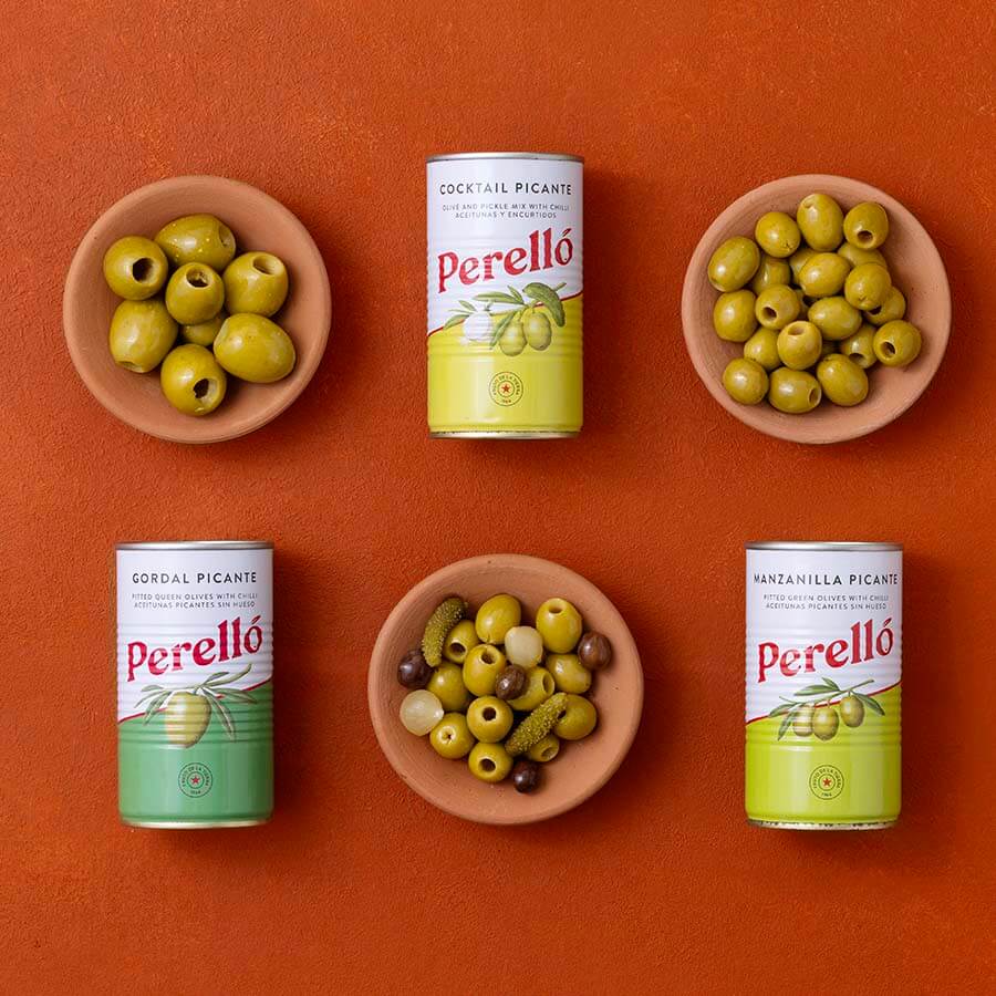 Four Perello tins, gordal, manzanilla and cocktail mix arranged on a terracotta background, with three olive dishes and one mixed olives-and-pickle dish placed between and around the tins.