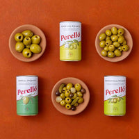 Four Perello tins, gordal, manzanilla and cocktail mix arranged on a terracotta background, with three olive dishes and one mixed olives-and-pickle dish placed between and around the tins.
