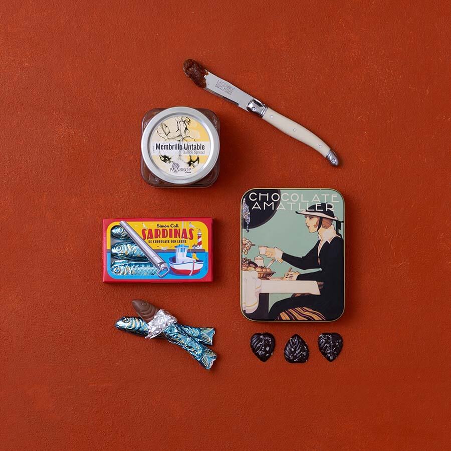 A styled flat-lay of Spanish food products: a jar of membrillo, a tin of chocolate leaves and a box of chocolate sardines.