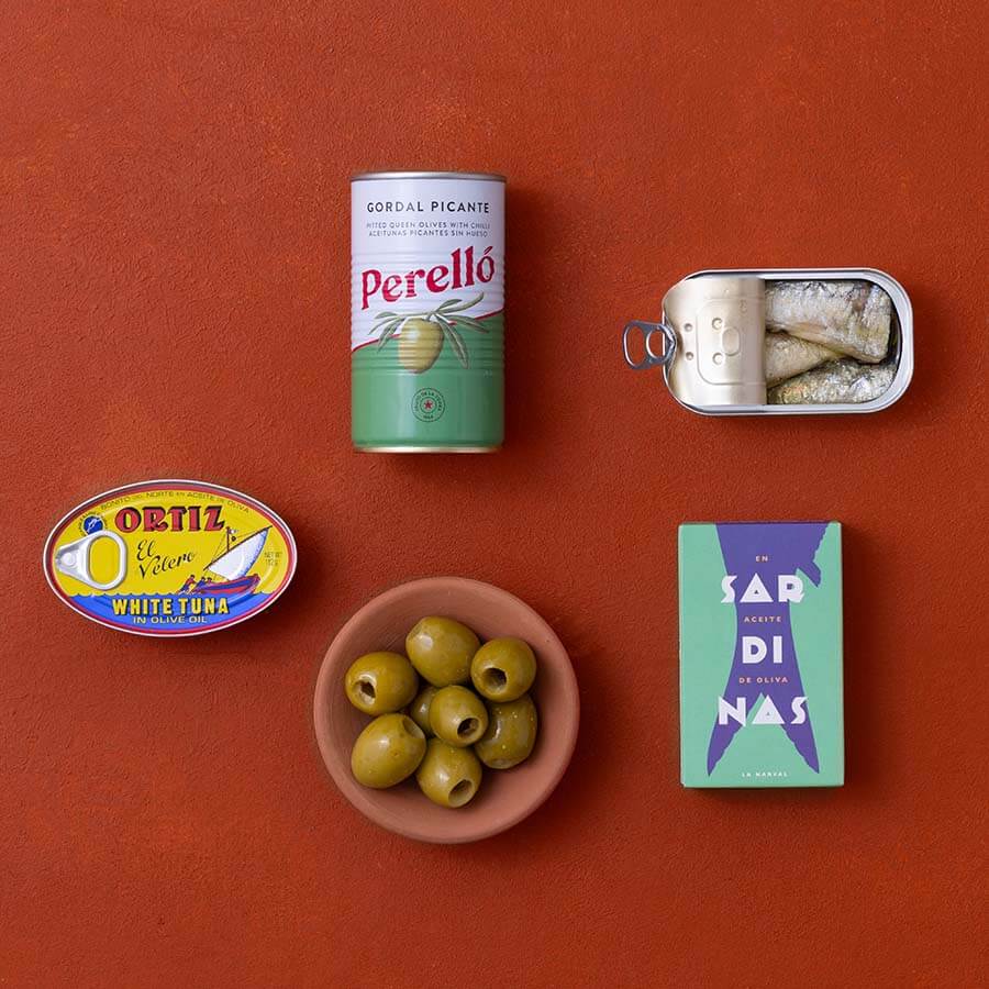 A styled flat-lay of food items on a terracotta background: a can of Perello gordal picante, a tin of sardines, an Ortiz white tuna tin and a bowl of green olives. 